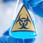 lab technician in gloves holding blue chemical in beaker with toxicity symbol